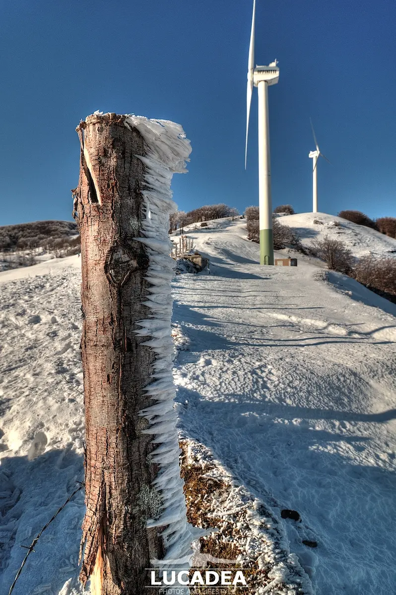 Frozen snow clinging to a shaded pole at Centocroci Pass, with wind turbines rising in the winter mountain landscape