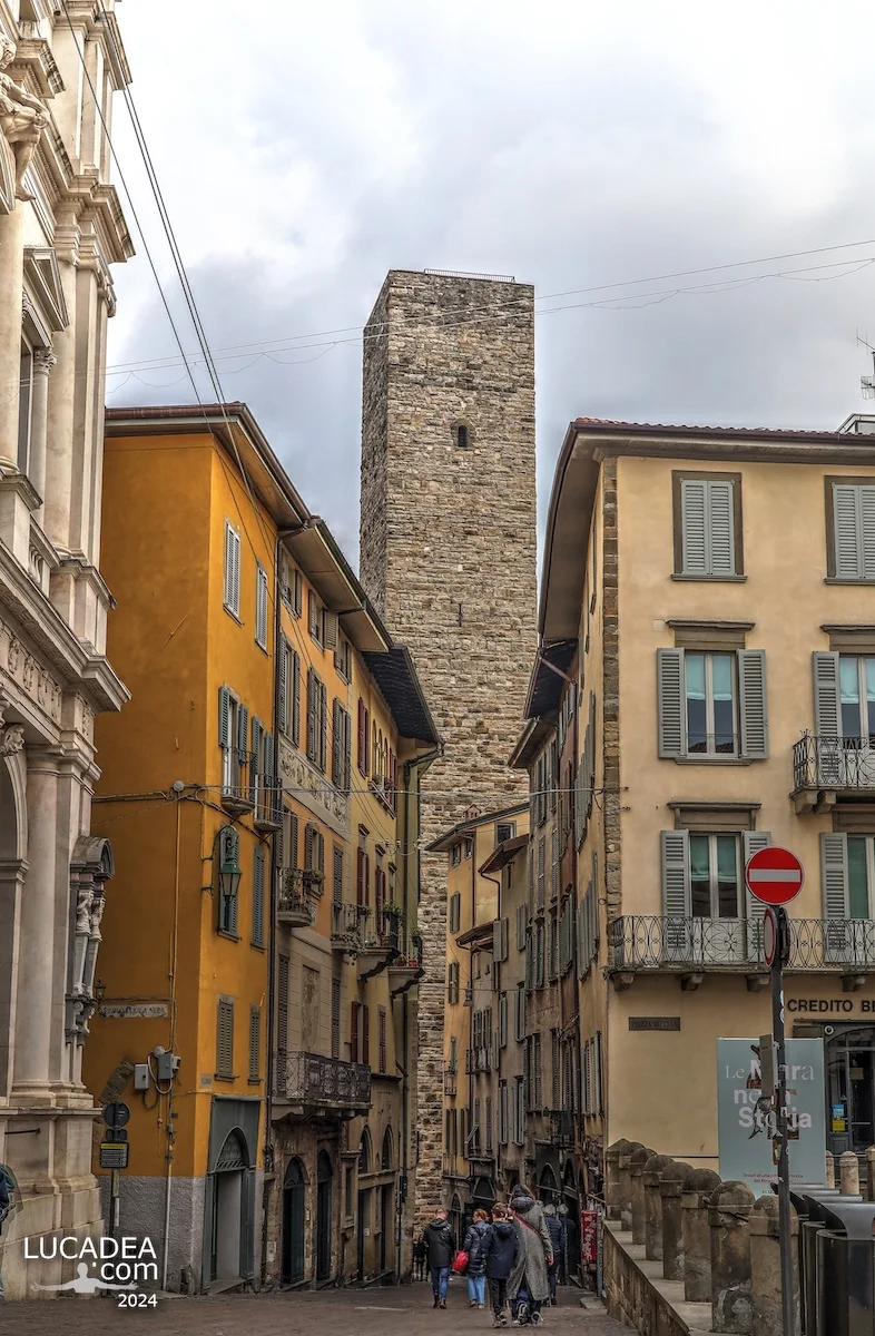 Vista della Torre del Gombito a Bergamo Alta tra edifici colorati, strada in discesa e persone che camminano