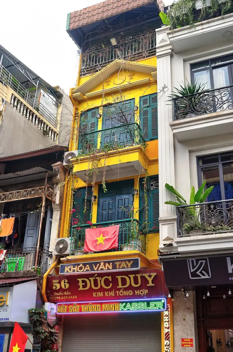 casa gialla Hanoi centro storico facciata stretta con balconi persiane verdi bandiera Vietnam piante rampicanti edifici vicini contesto urbano
